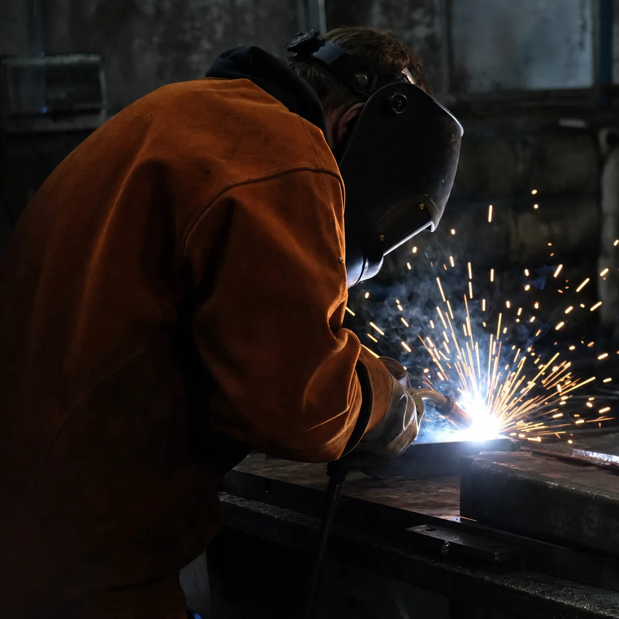 A fabricator in the rust-orange jacket striking an arc, sparks arcing off the workpiece in a dim shop.
